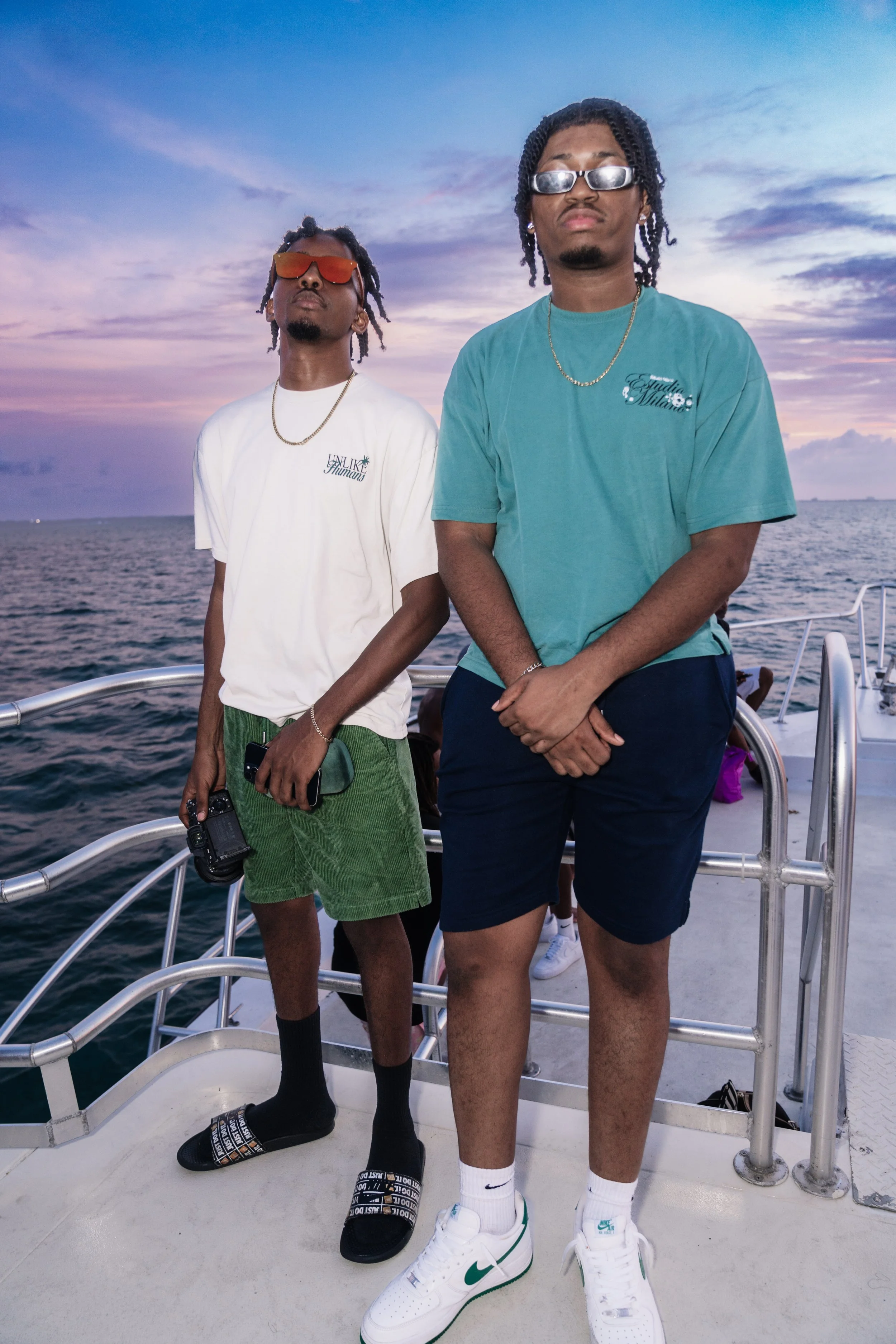 Two young men standing on the deck of a boat during sunset, with the ocean and sky in the background. They are wearing casual summer clothing, sunglasses, and jewelry, posing confidently for the photo.