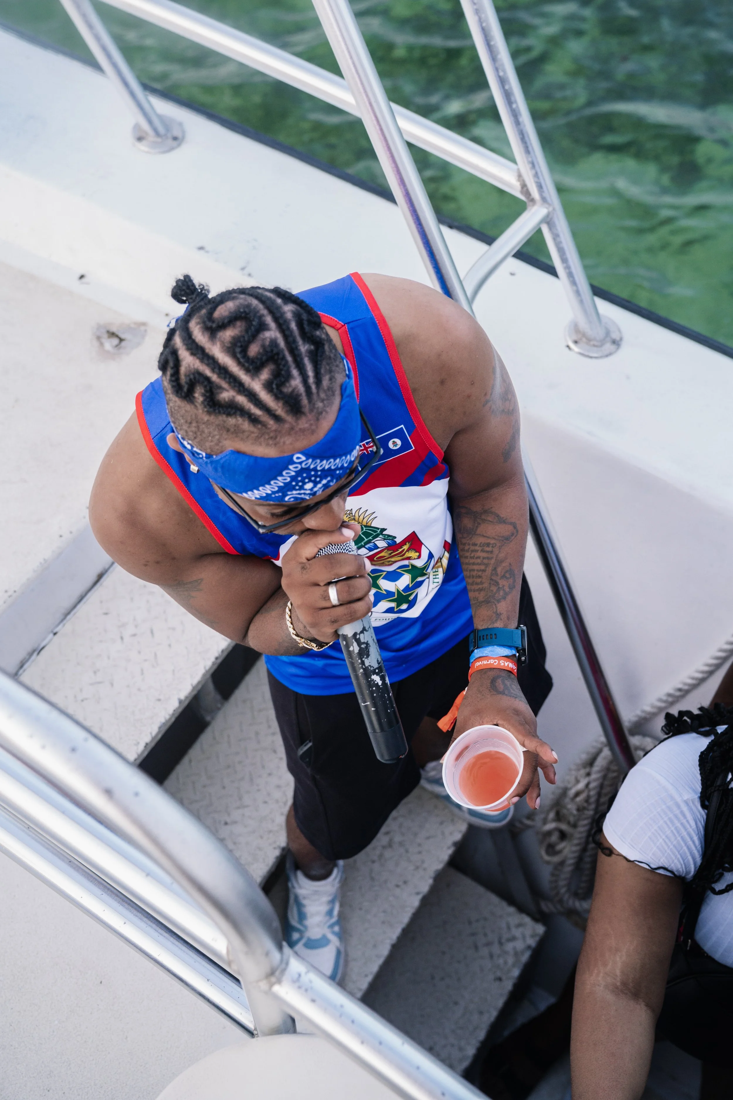 Person with braided hair, wearing a blue bandana and athletic attire, holding a microphone and a cup of pink drink, standing on boat stairs.