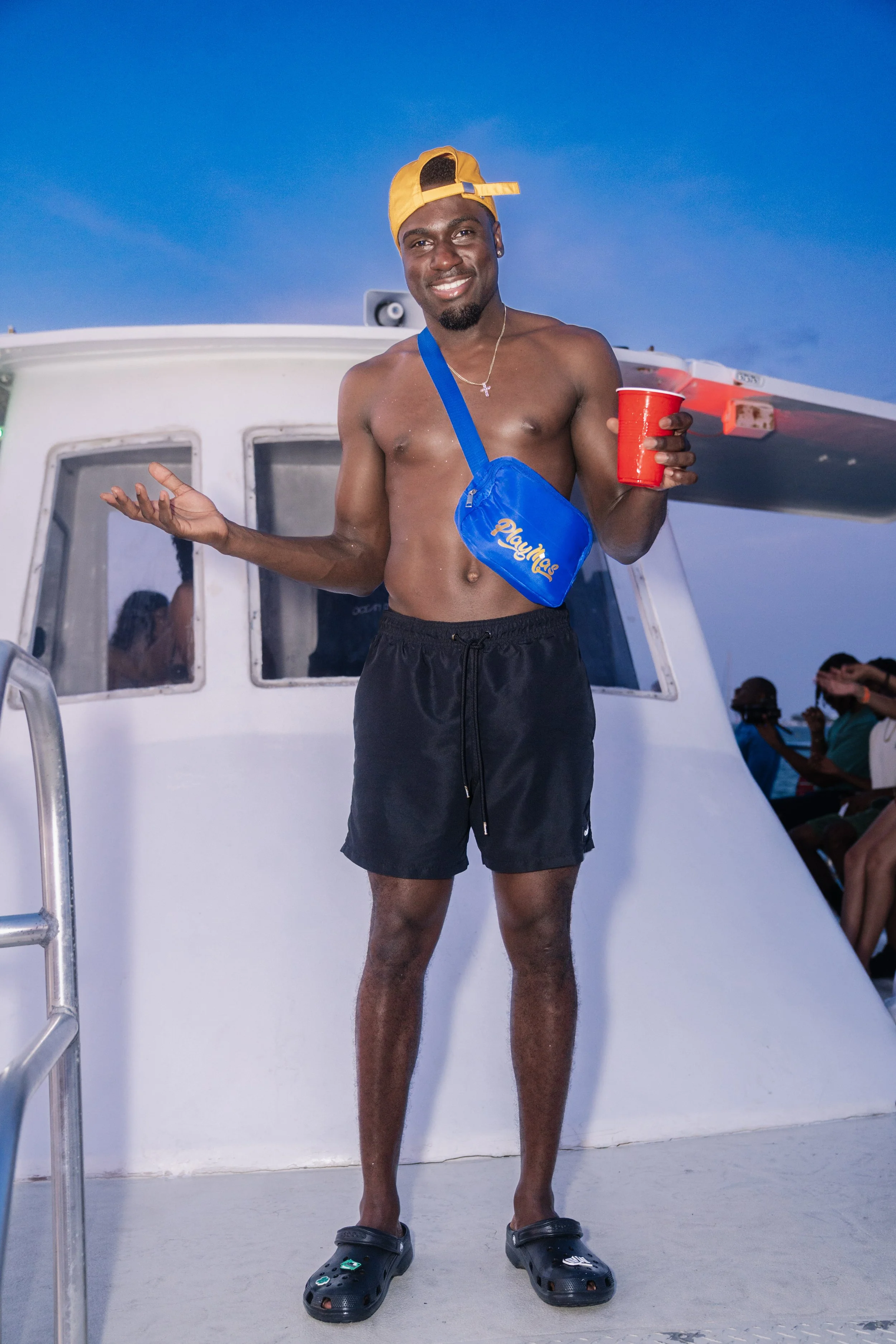 A shirtless young man wearing a blue crossbody bag, black shorts, and black Crocs, holding a red cup, standing on the deck of a boat with a blue sky background. He is smiling and wearing a yellow backwards cap.