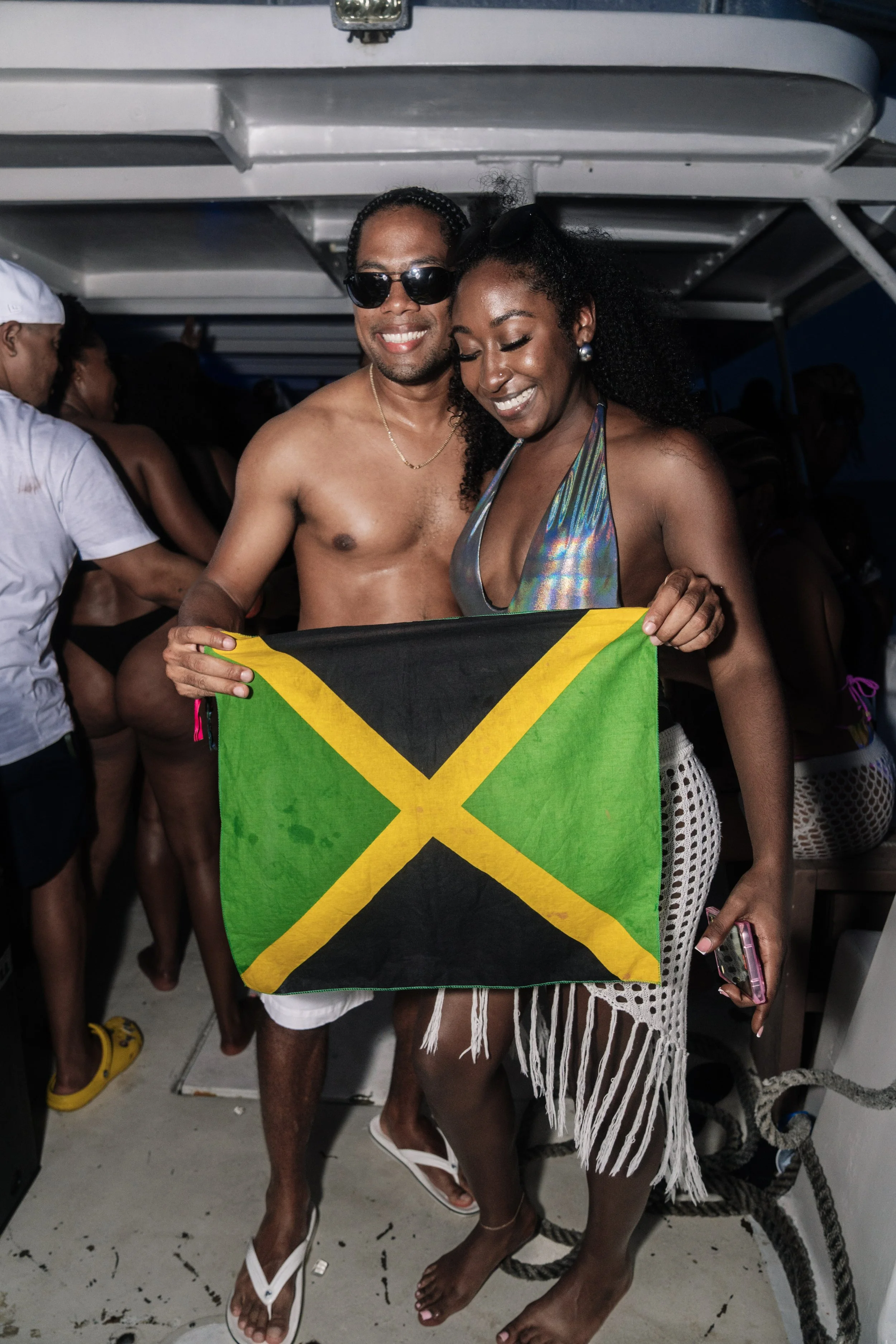Two people on a boat holding a Jamaican flag, surrounded by other party-goers in swimwear.
