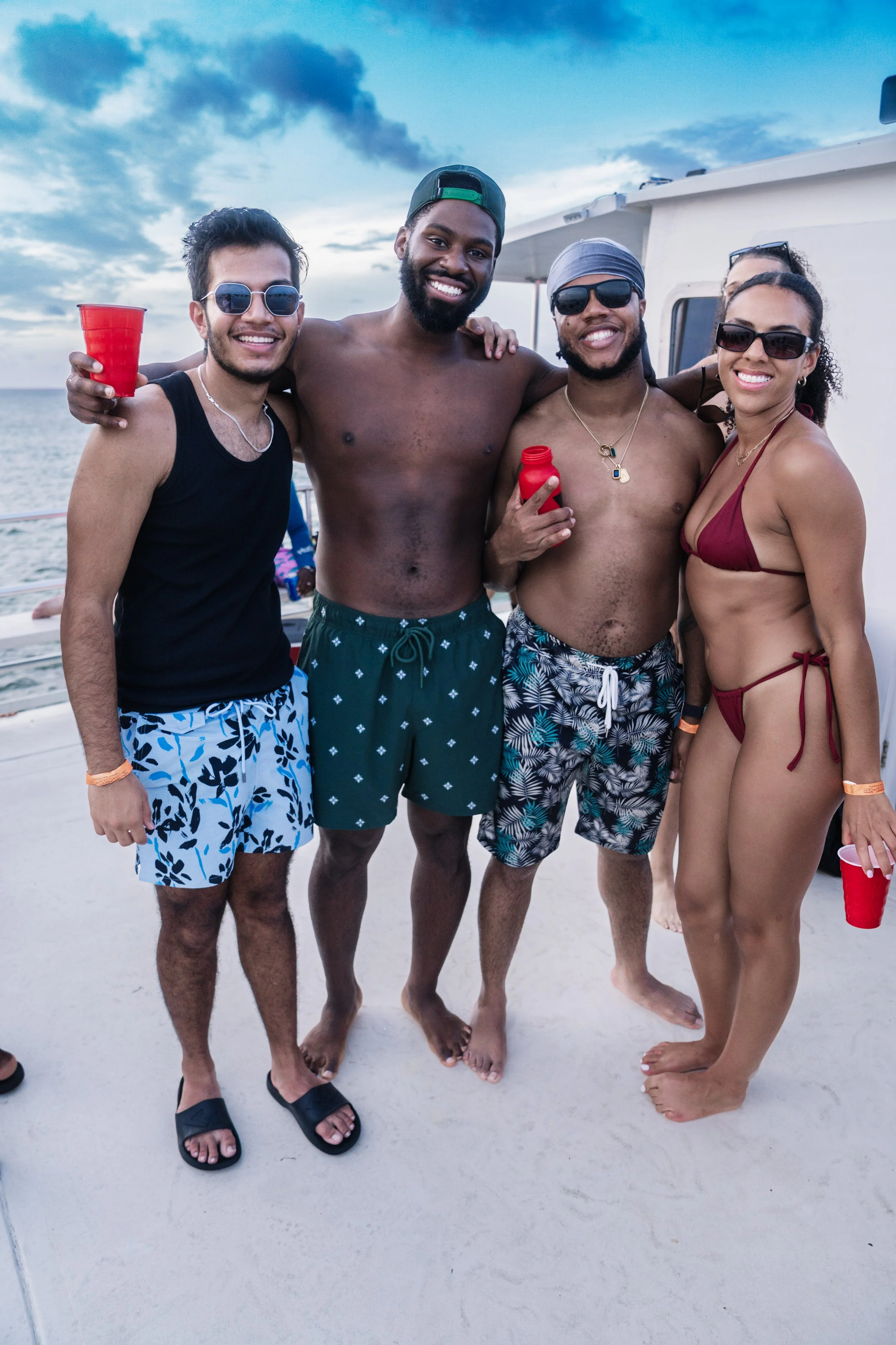Group of five friends enjoying a boat party on the beach, wearing swimwear and sunglasses, holding red cups, with the ocean and partly cloudy sky in the background.