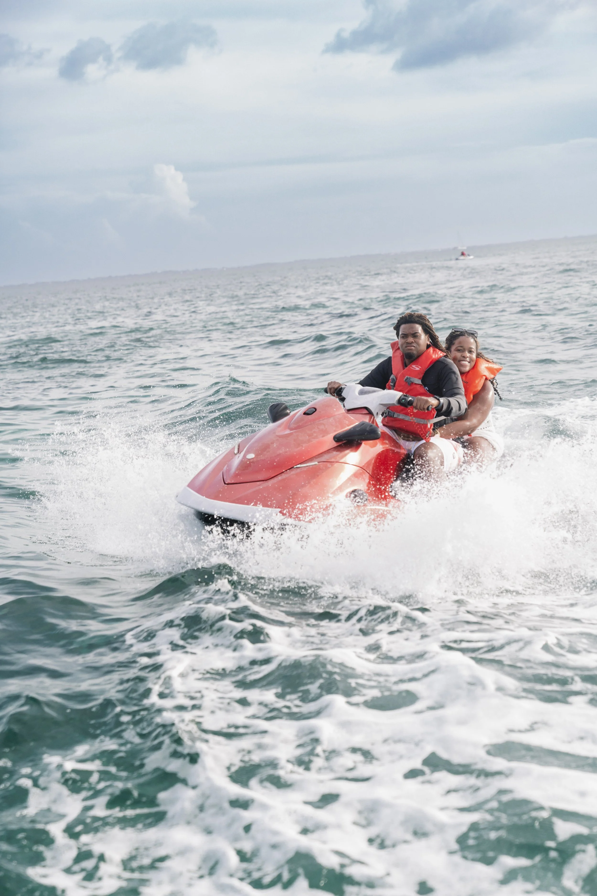 Two people riding a red jetski on the water, wearing life jackets and smiling, with a cloudy sky in the background.