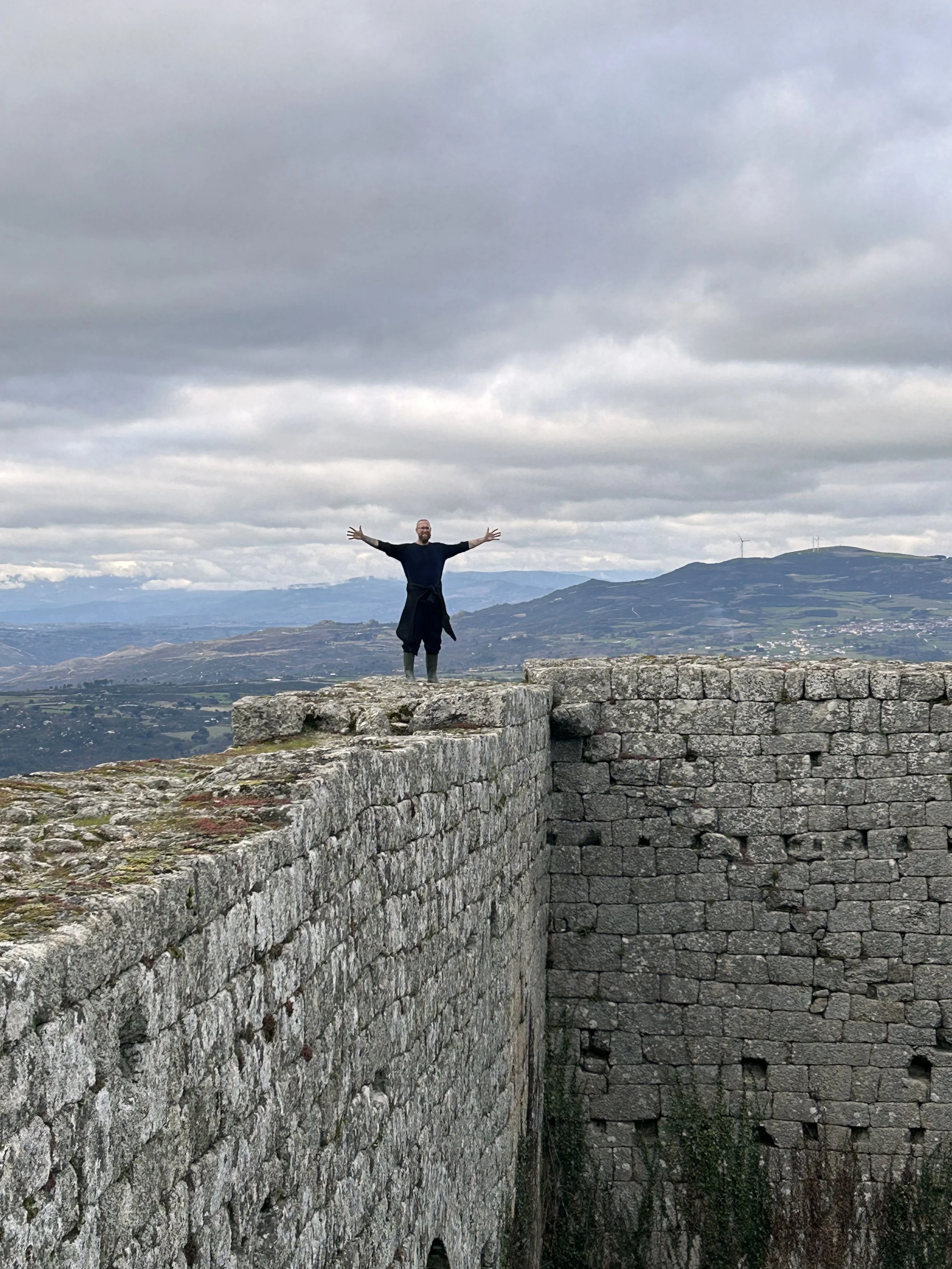 Billy Mills standing on a stone fortress wall with arms outstretched, overlooking rolling hills under a cloudy sky.