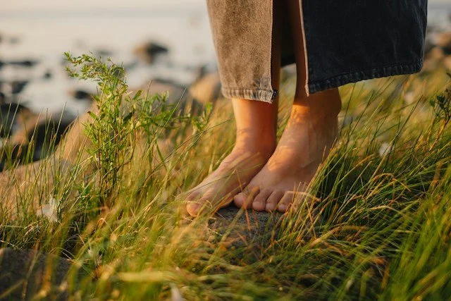 Bare feet standing on grass and stones near a body of water, with long fabric draping over the ankles.