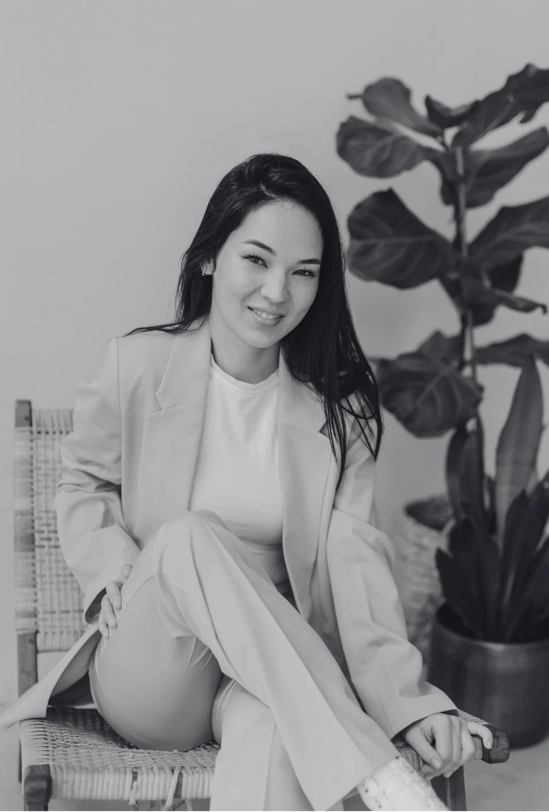 Black and white photo of a person sitting on a chair wearing a suit, with a plant in the background.