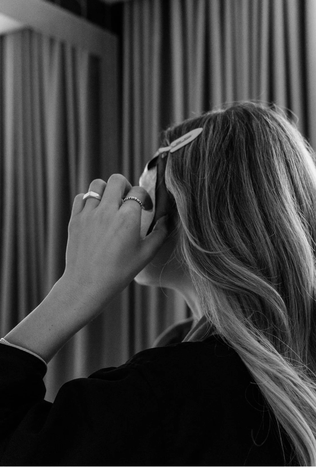 Black and white image of a woman with long hair adjusting sunglasses, wearing rings, in front of curtains.