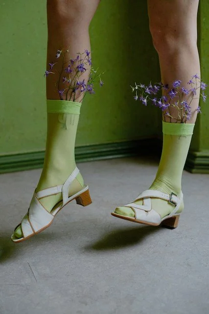 Person wearing light green socks with purple flowers inserted, styled with white sandals against a green background.