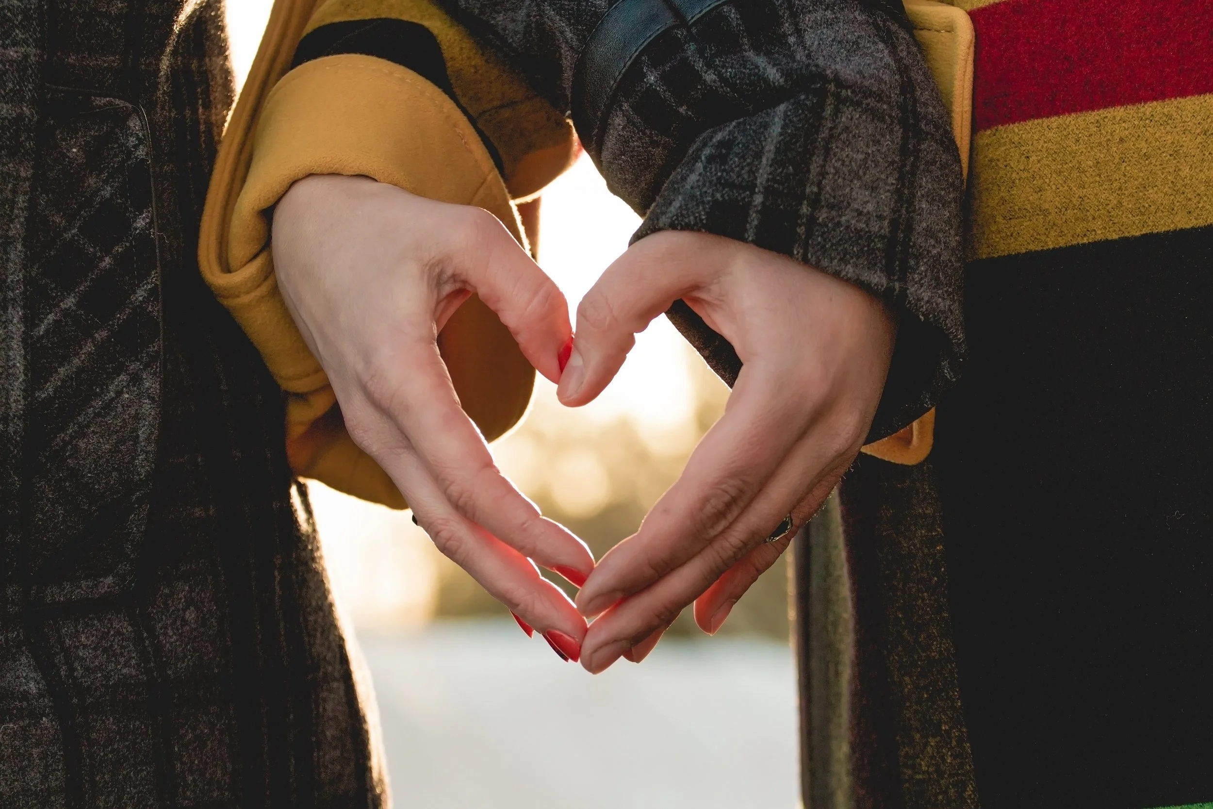 Two people forming a heart shape with their hands in front of a bright background.