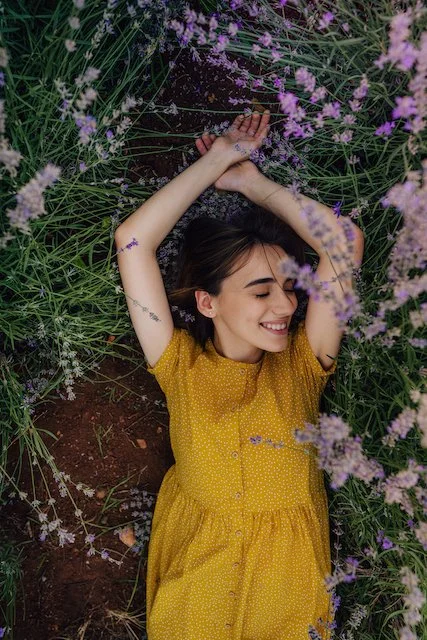 Woman in yellow dress lying in lavender field, smiling with eyes closed, surrounded by flowers.