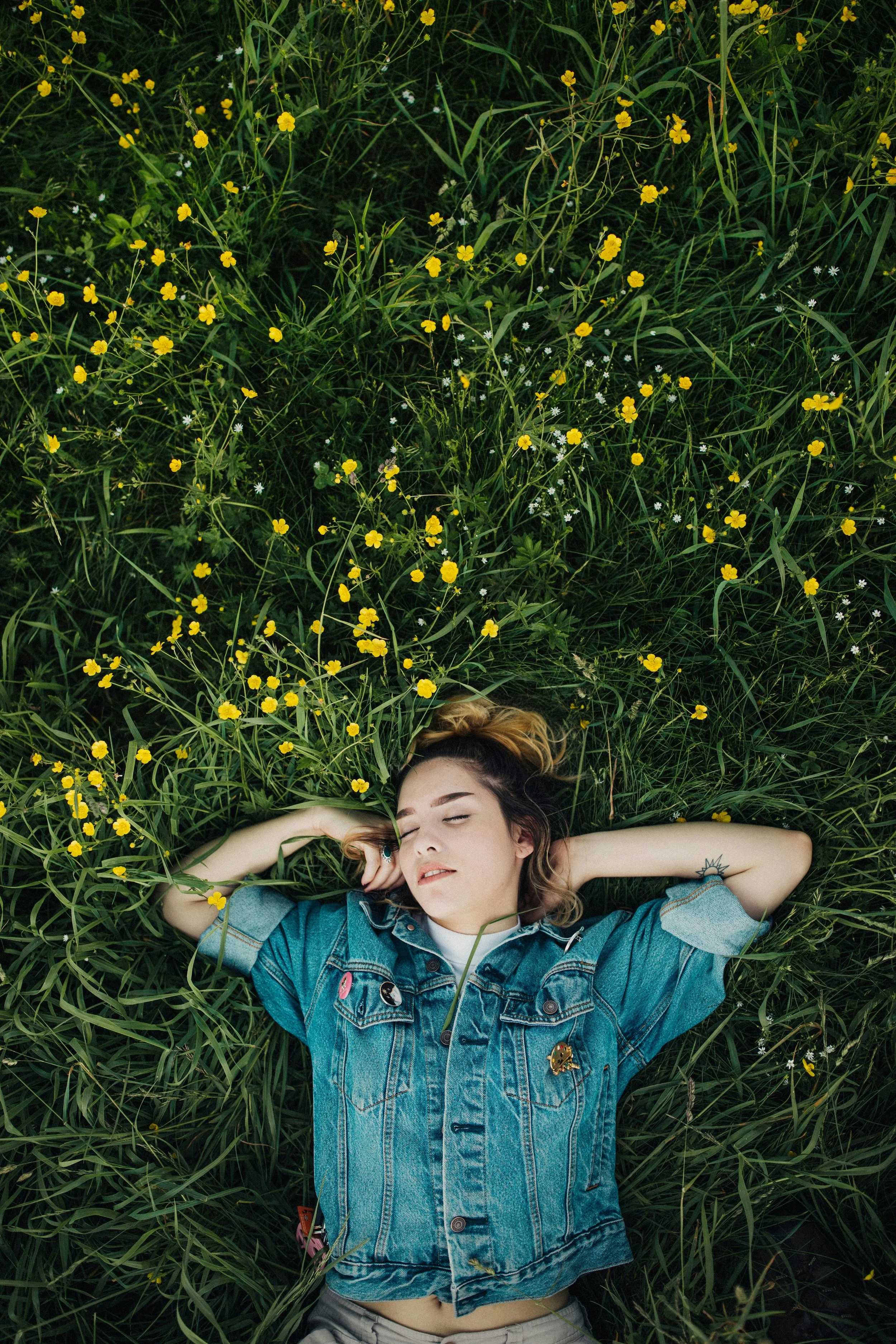 Person wearing a denim jacket lying on grass surrounded by yellow flowers.