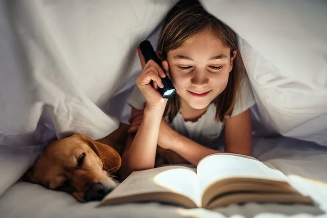 Girl reading a book under a blanket with a flashlight, accompanied by a sleeping dog.
