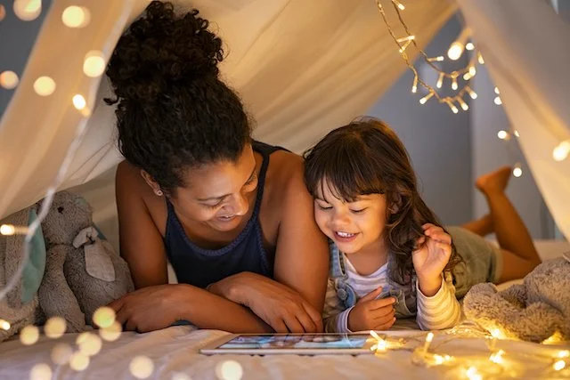 Mother and daughter lying under a blanket fort, smiling and looking at a tablet, surrounded by fairy lights.