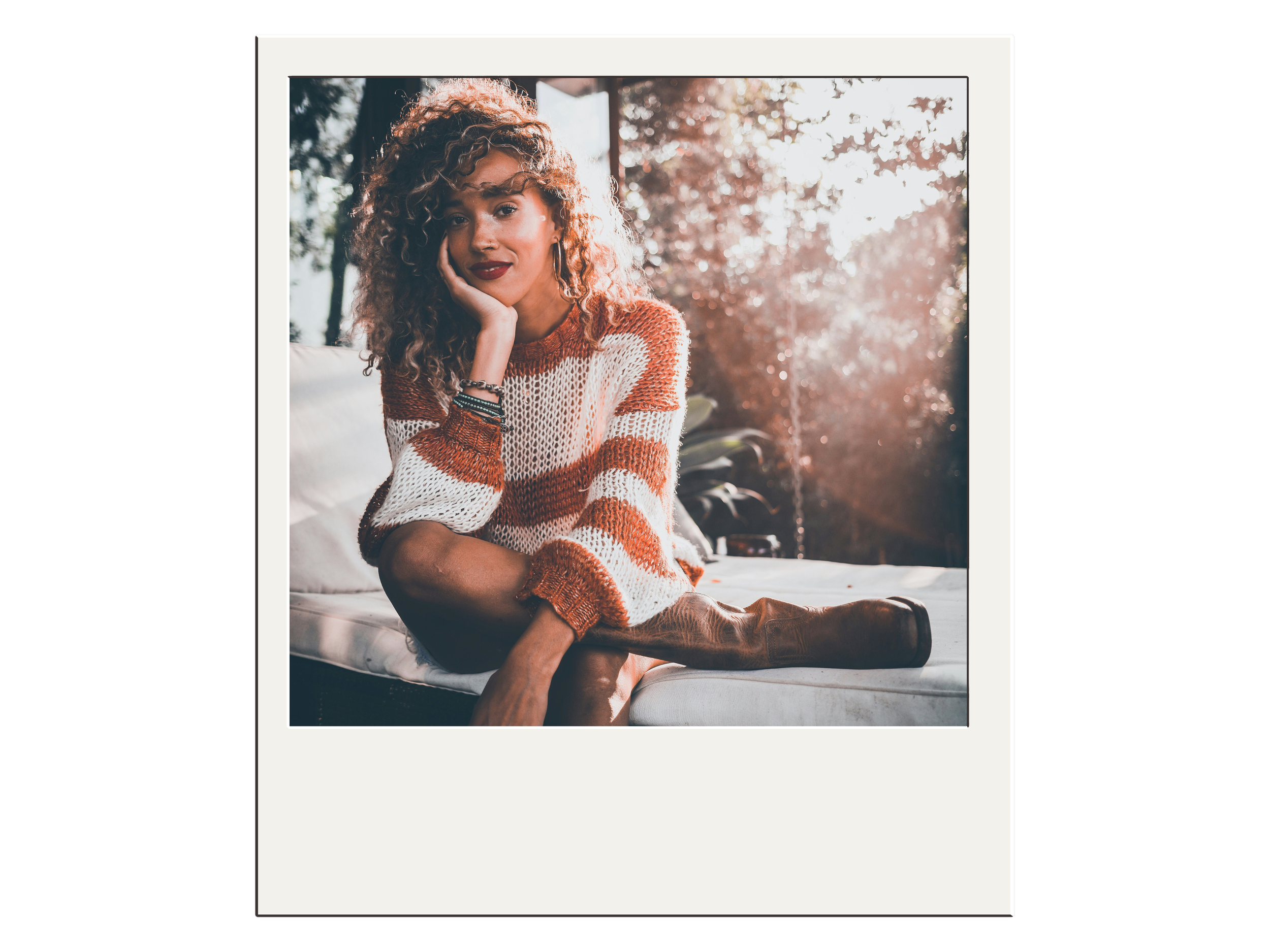 Woman sitting outdoors wearing striped sweater and boots, with hand on cheek, trees in background.