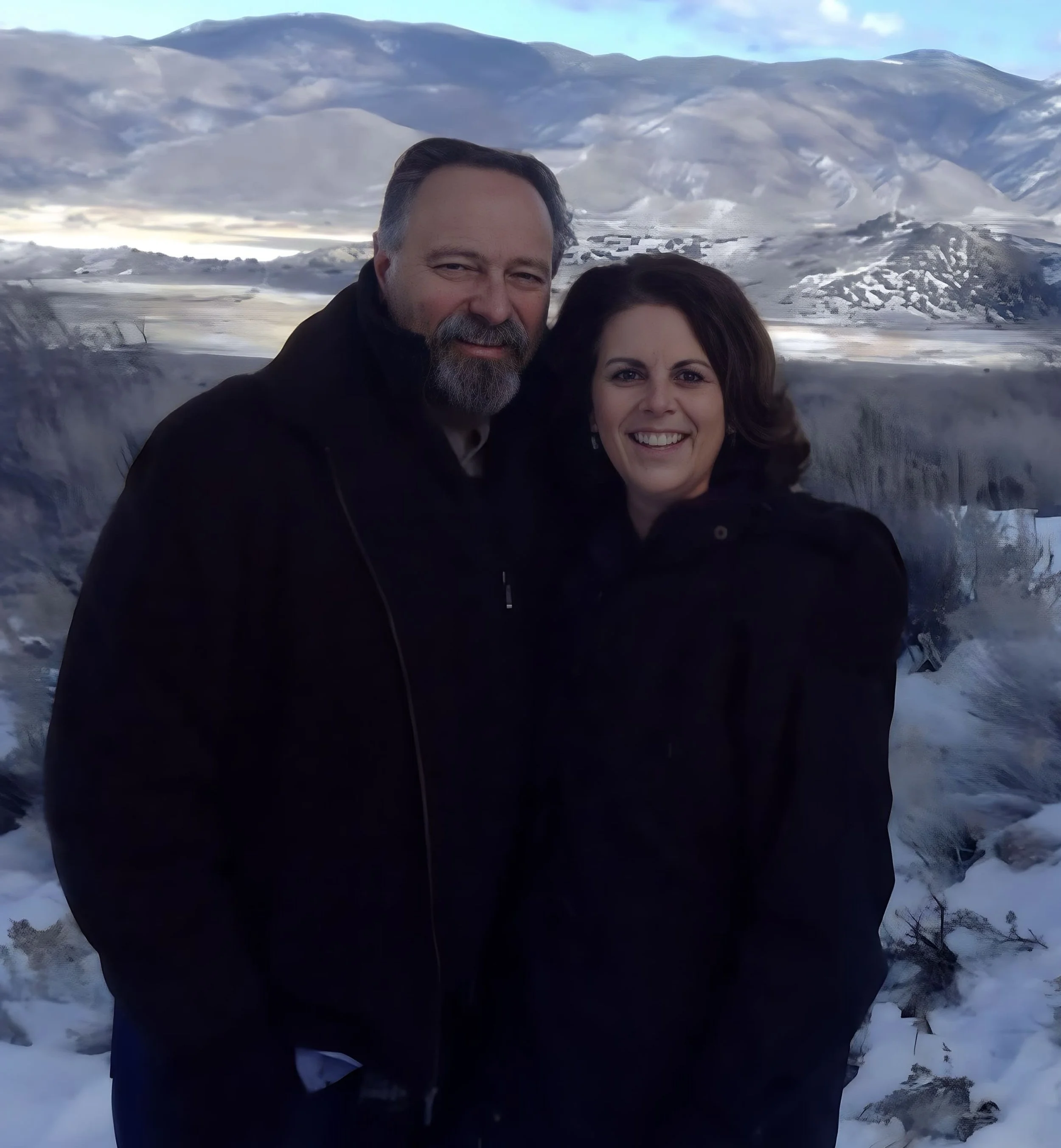 A man and woman smiling in front of a snowy landscape with mountains and a waterfall.