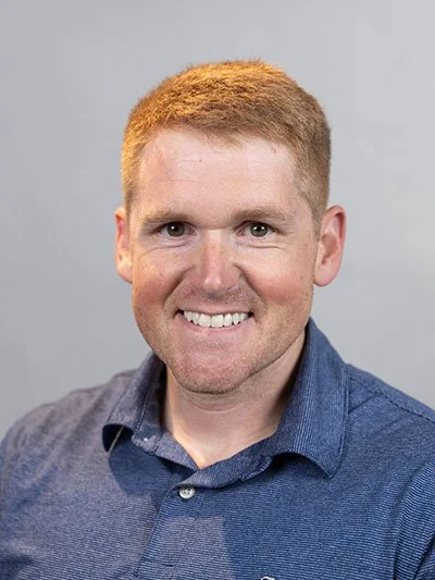 Headshot of a smiling man with red hair and a beard, wearing a blue collared shirt against a gray background.