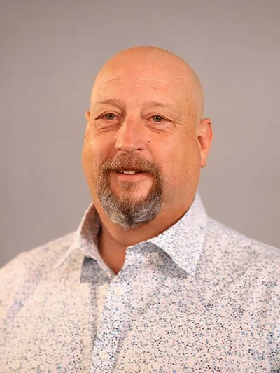 A portrait of a middle-aged man with a bald head, goatee, and mustache, wearing a white button-up shirt with a floral pattern, against a neutral background.