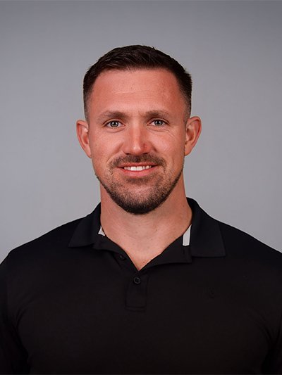 Headshot of a man with short dark hair, a beard, wearing a black collared shirt, against a gray background.
