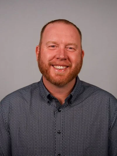 Headshot of a smiling man with a beard and short hair, wearing a patterned button-up shirt against a plain gray background.