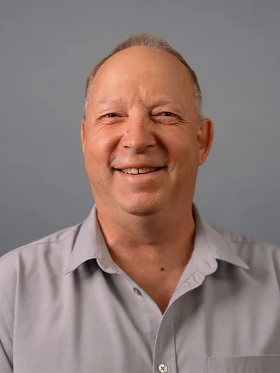 A smiling middle-aged man with short gray hair wearing a light gray collared shirt, standing against a plain gray background.