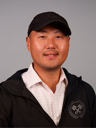 A man wearing a black cap, black jacket, and a white collared shirt, smiling against a gray background.