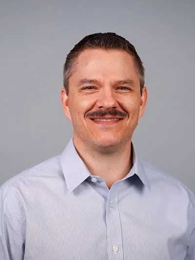 Portrait of a man with short dark hair, a mustache, wearing a light blue collared shirt, smiling against a plain gray background.