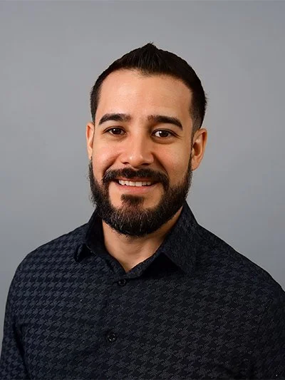 A man with dark hair and a beard, wearing a black patterned shirt, smiling against a gray background.