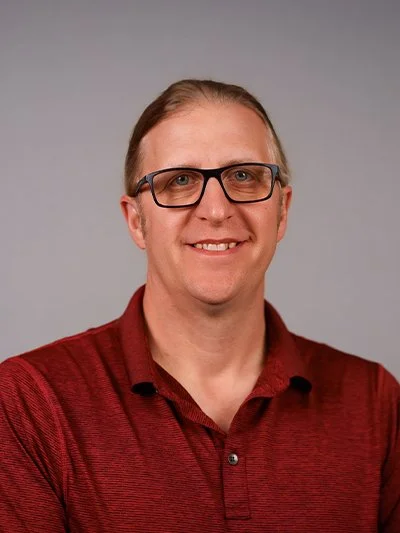 Portrait of a man wearing glasses and a red polo shirt, smiling against a plain gray background.