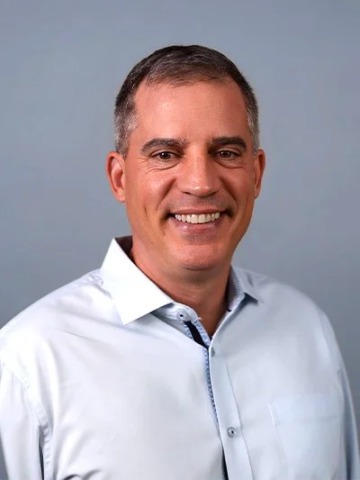 A man with short dark hair, wearing a light blue dress shirt, smiling against a plain gray background.