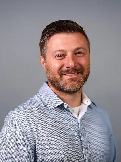 A smiling man with dark hair and a beard, wearing a light blue button-up shirt, standing against a plain gray background.
