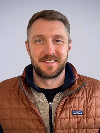 A man with brown hair and a beard smiling, wearing a brown Patagonia jacket and a black shirt, against a plain light gray background.