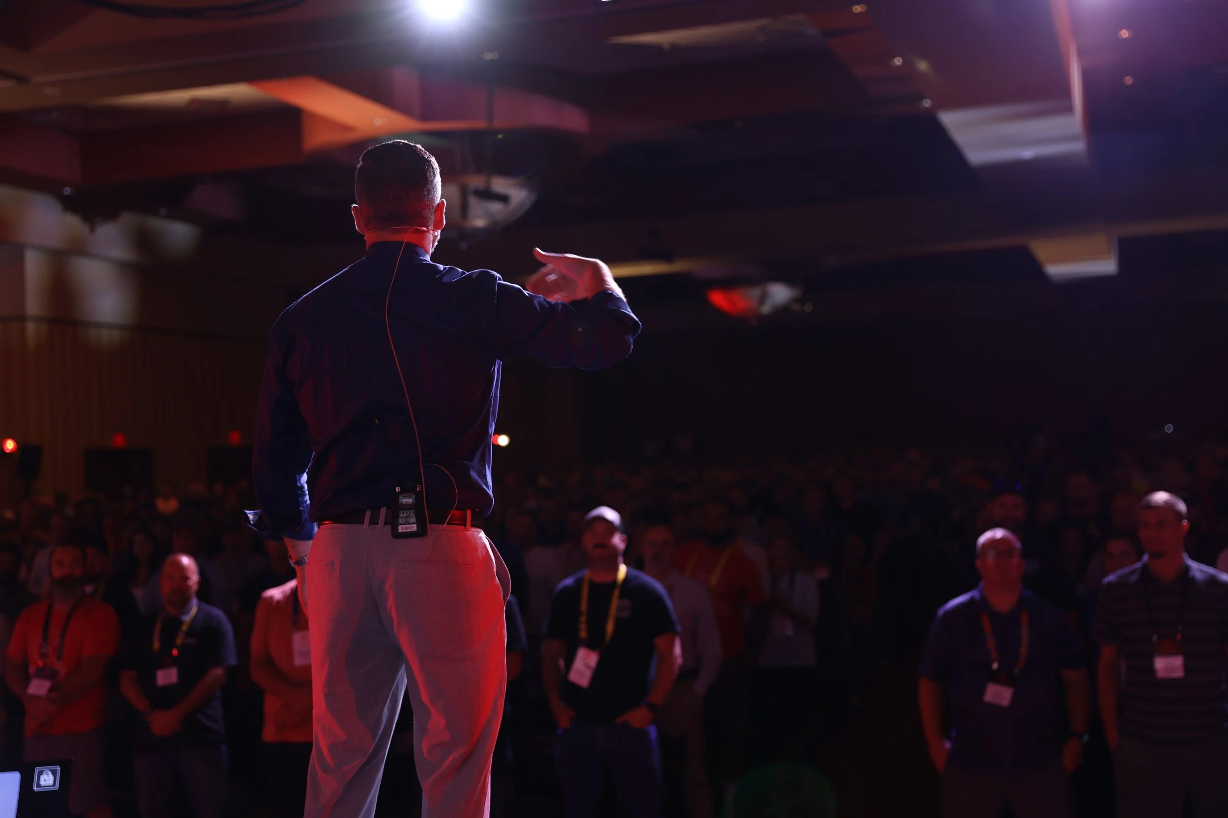 A man in a dark shirt and light-colored pants speaking or performing on stage at a conference or event, with a large audience watching.