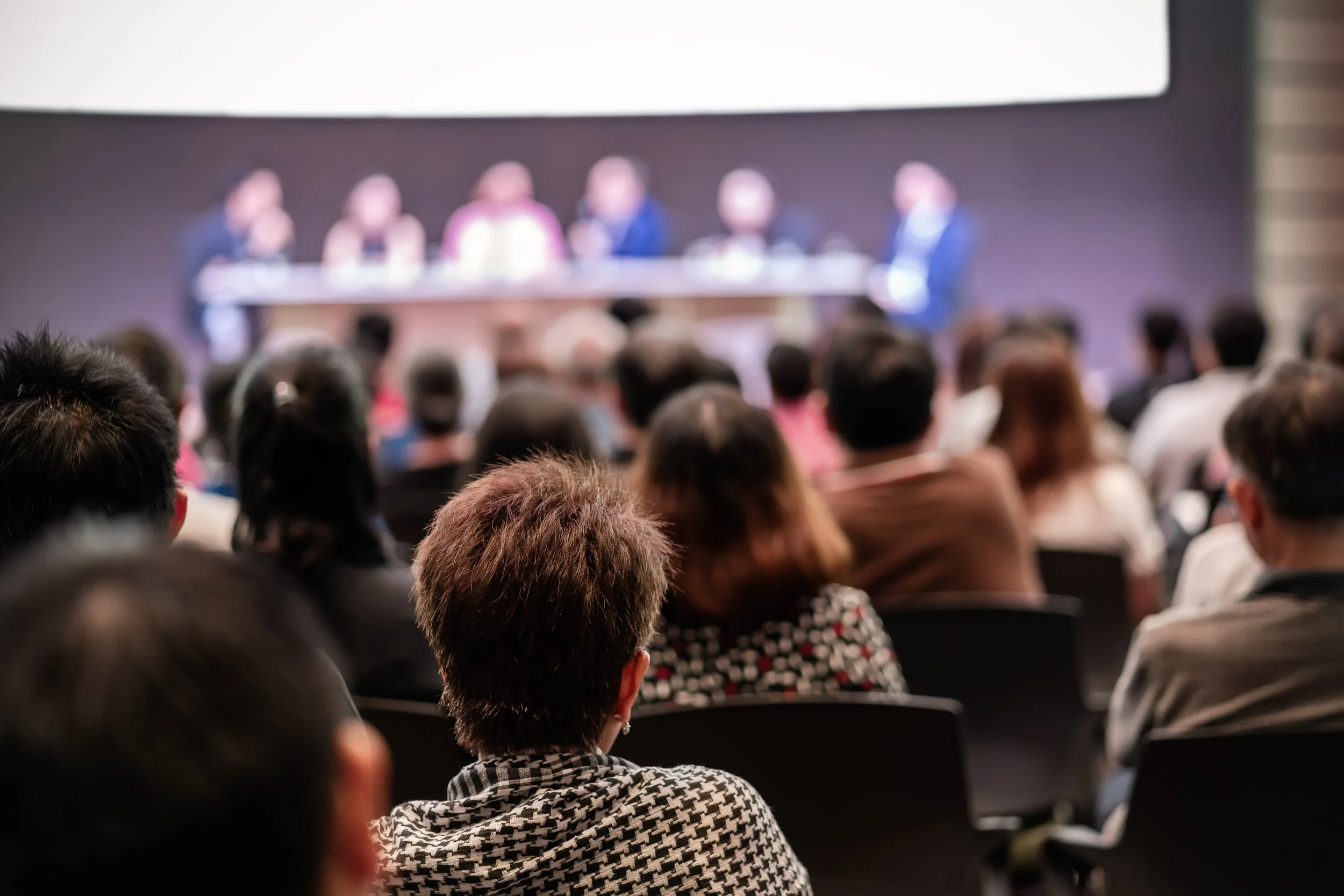 An audience attending a conference or panel discussion, with several speakers or panelists blurred on a stage in the background.