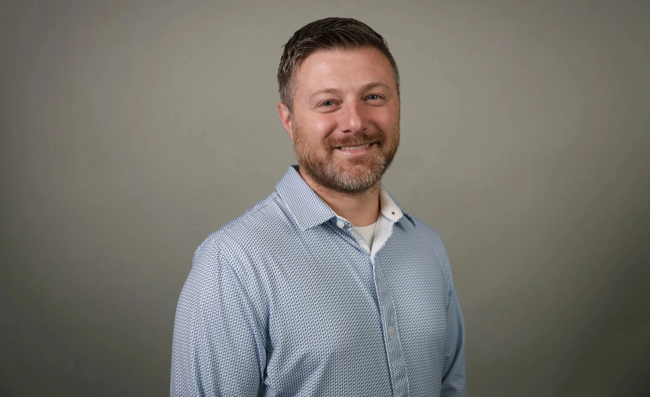 Portrait of a smiling man with short brown hair and beard, wearing a light blue patterned dress shirt against a plain gray background.