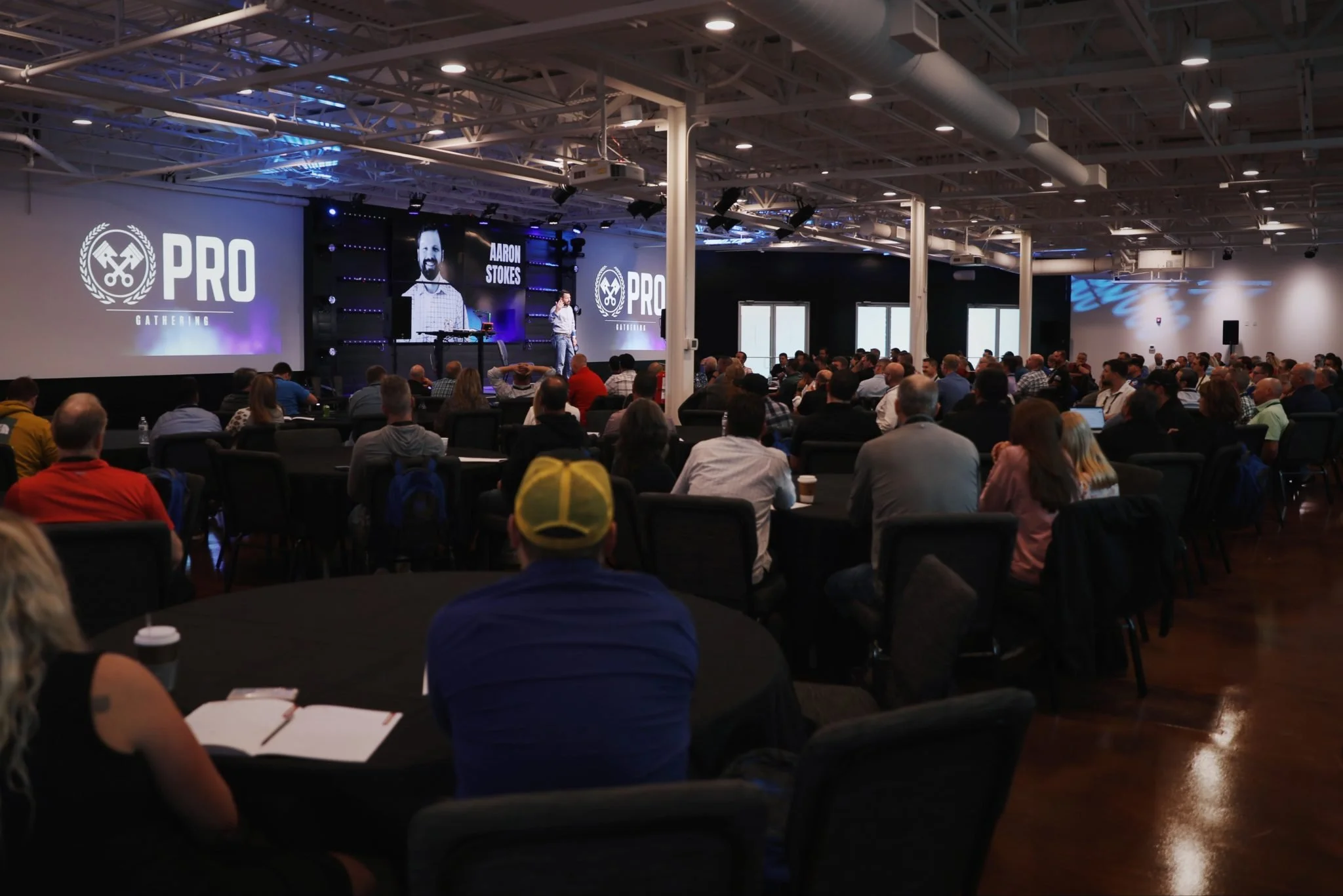 Large conference room filled with seated audience attending a presentation; on stage, a speaker has a large screen behind him displaying event branding and a photo of a man.