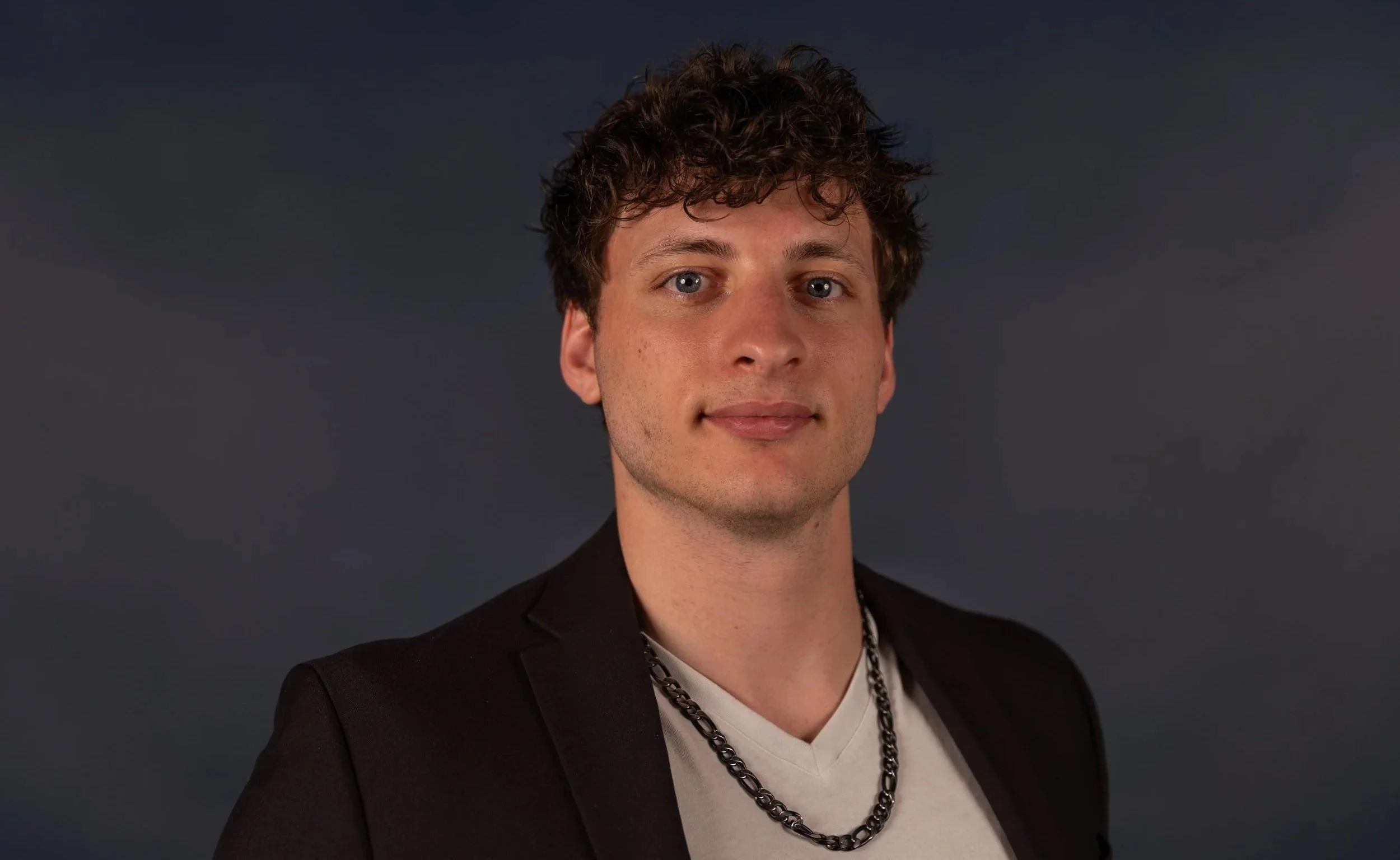 A young man with short, curly red hair and fair skin wearing a blue collared shirt, standing against a plain gray background.