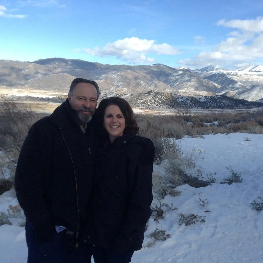 A smiling couple standing outdoors in a snowy landscape with mountains in the background.