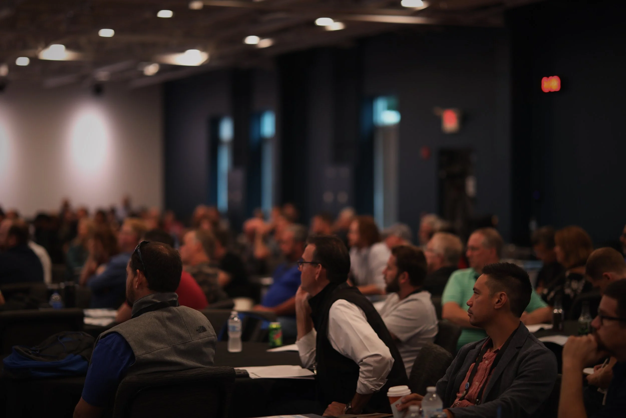 A large group of people sitting and listening during a conference or seminar in a dimly lit room with dark walls and ceiling.
