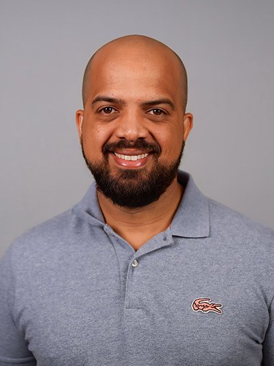 A smiling man with a bald head, beard, and mustache, wearing a light blue polo shirt with a small crocodile logo on the chest, against a plain gray background.