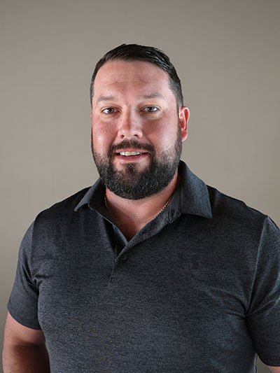 Portrait of a man with short dark hair, a full beard, wearing a dark gray polo shirt, smiling against a plain beige background.