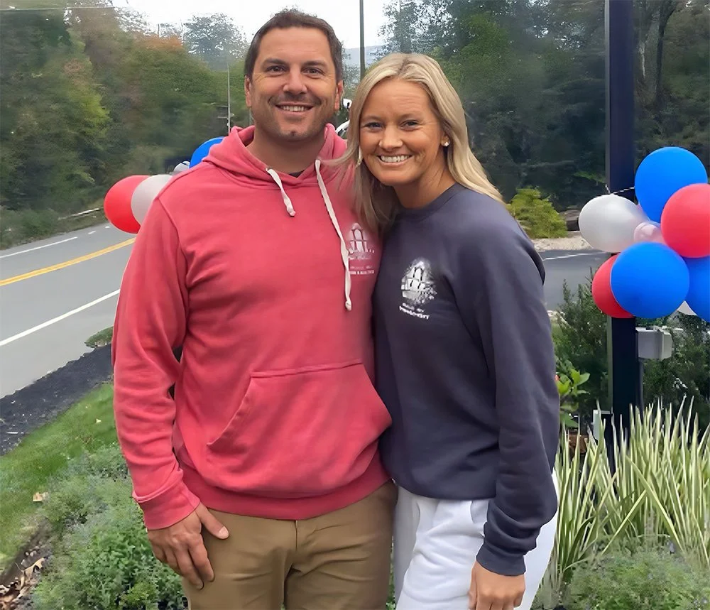 A man and woman smiling outdoors, standing next to a glass window decorated with balloons in red, white, and blue, with trees and a road visible in the background.