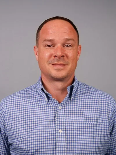 Headshot of a man with short dark hair, light skin, wearing a blue and white checkered button-up shirt, against a plain gray background.