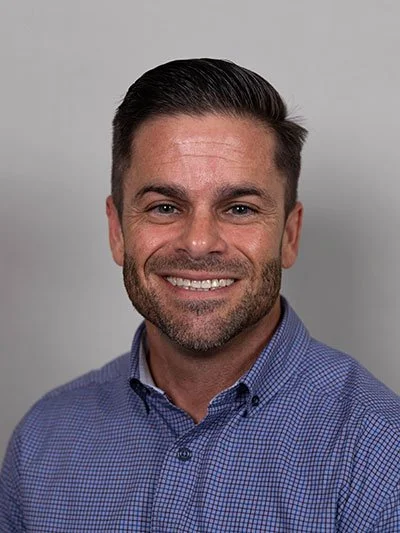 A smiling man with short dark hair and a beard, wearing a blue checkered button-up shirt, against a gray background.