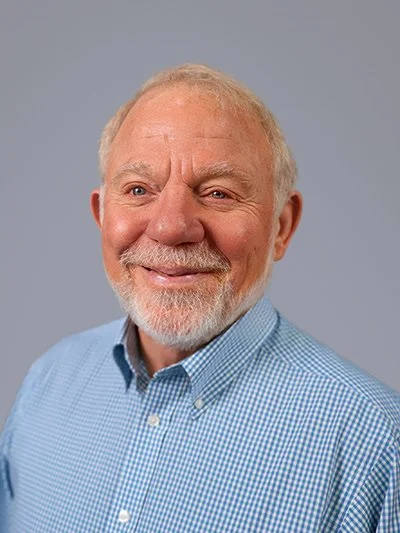Portrait of a smiling elderly man with a beard, wearing a light blue checkered shirt against a gray background.