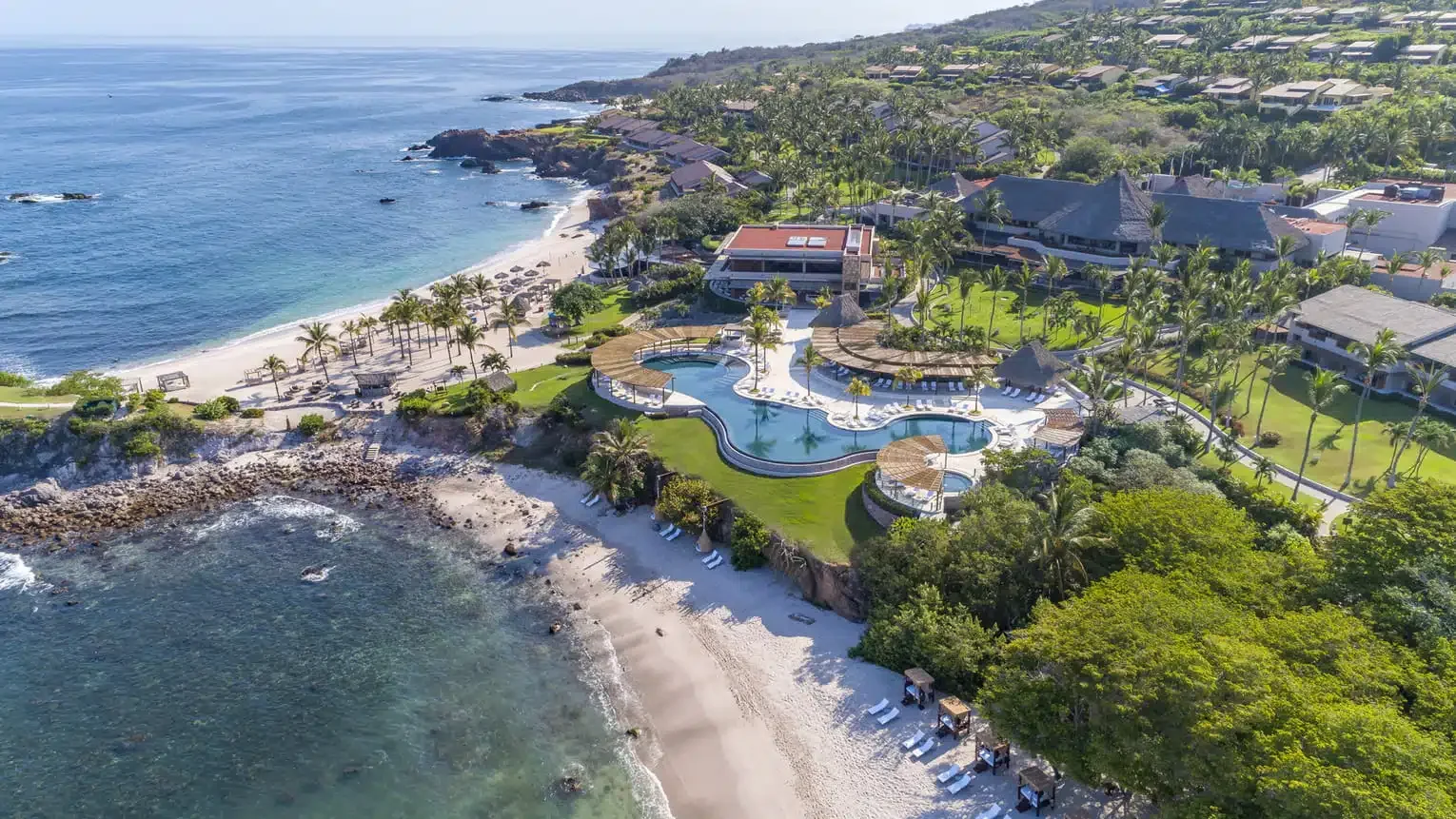Aerial view of a luxurious beachfront resort with a large lagoon-style swimming pool, surrounded by palm trees, beach chairs, and cabanas, overlooking the ocean with a rocky shoreline.