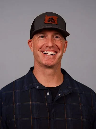 A smiling man wearing a gray and black trucker hat with an orange patch and a dark plaid shirt against a plain gray background.