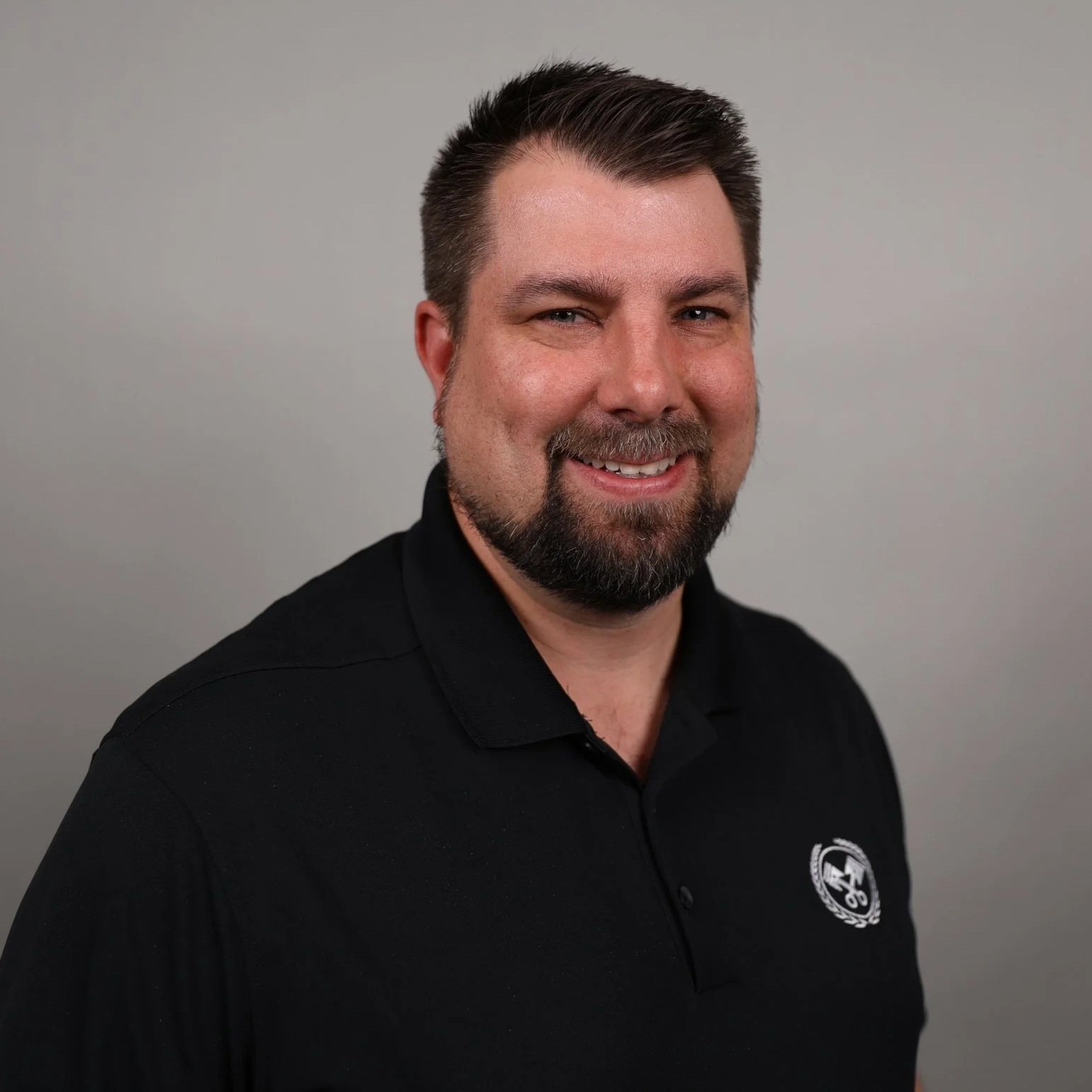 Portrait of a smiling man with short dark hair, beard, and mustache, wearing a black polo shirt against a plain, light gray background.