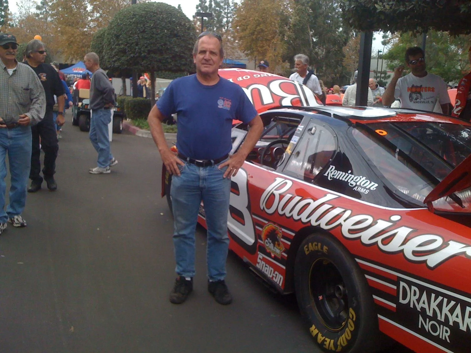 A man standing next to a red race car at an outdoor event with several people in the background.
