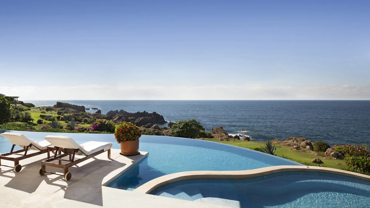 Luxury swimming pool overlooking the ocean with two lounge chairs and a potted flowering plant, surrounded by greenery and rocky coastline.