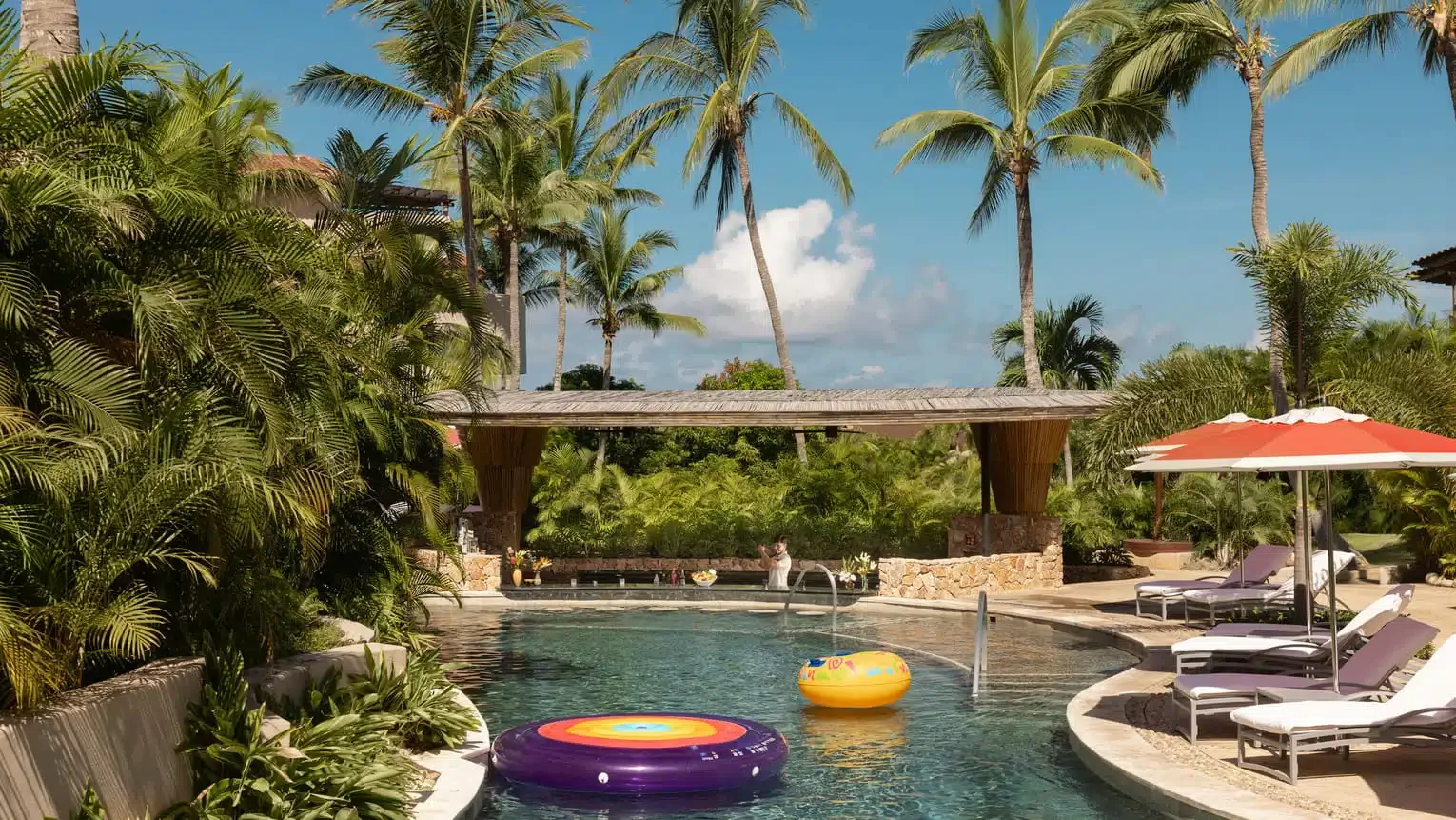 Swimming pool surrounded by tropical plants, lounging chairs with umbrellas, and tall palm trees under a blue sky with clouds.