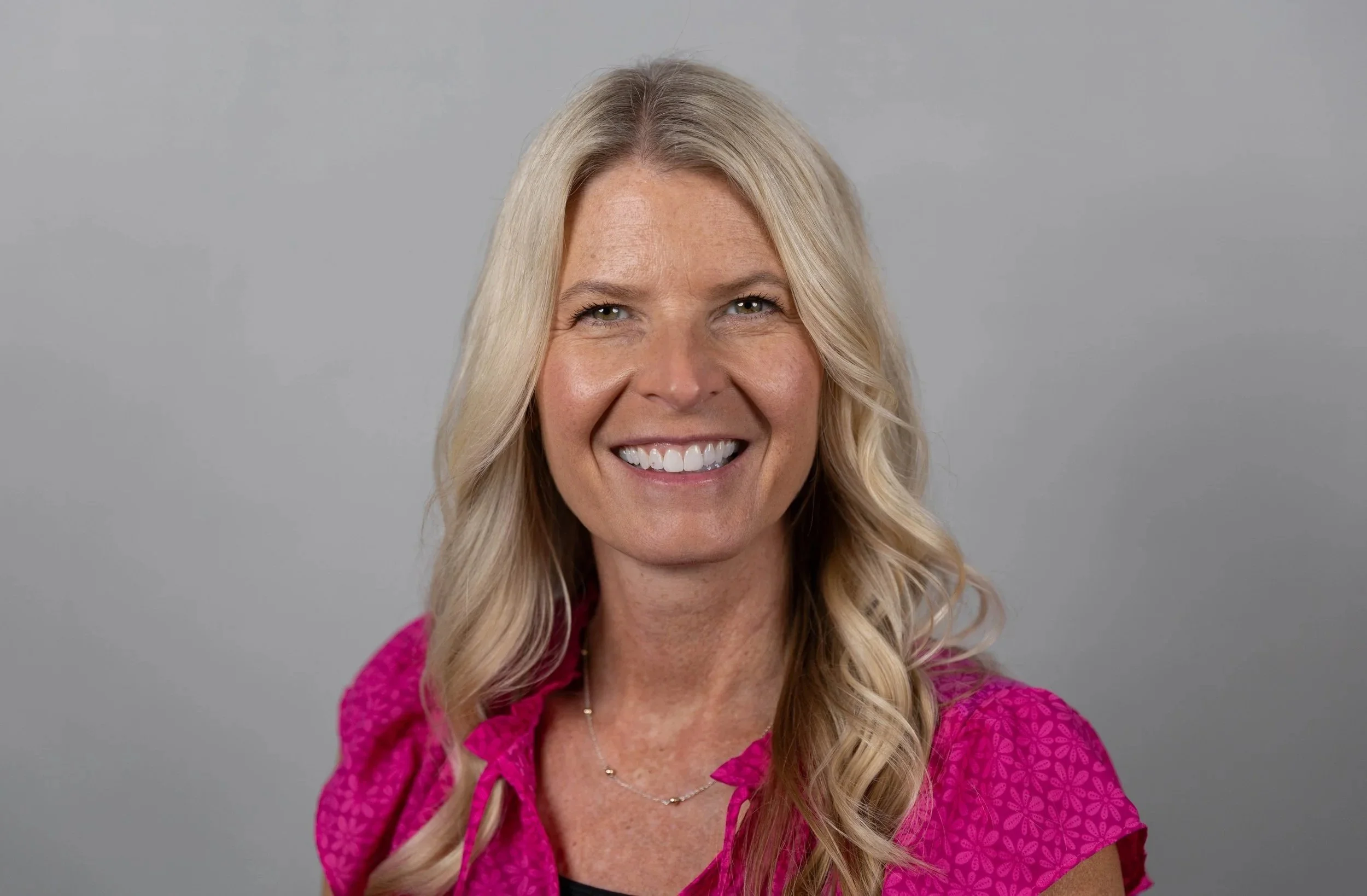 A woman with long blonde hair smiling, wearing a pink top with puffed sleeves, against a gray background.